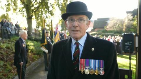 Lt Col Mark Jackson stood wearing a black bowler hat and black coat, with four army medals on display 