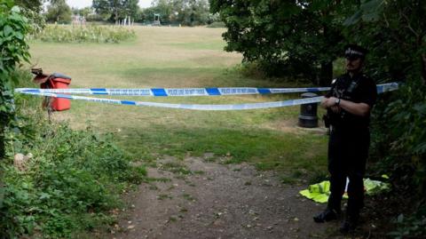 A park in Ipswich cordoned off with police tape. A police officer is also standing nearby. 