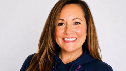 Head and shoulders of Sam Wright. She is smiling and has long straight brown hair and is wearing a dark blue zip up top