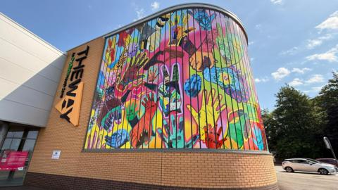 A large multi-coloured mural on a curved brick wall on a building, with lots of different patterned hands on it. There is a sign next to it saying "The Way".