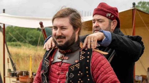 A man helping another man get dressed up as a knight, with a red tunic and black sleeveless top. Both are standing in front of a tent at a reenactment event.