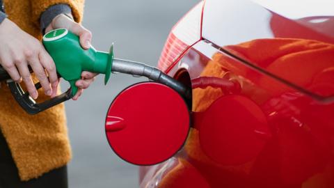 A woman in a yellow coat using a green fuel pump to refuel a red car