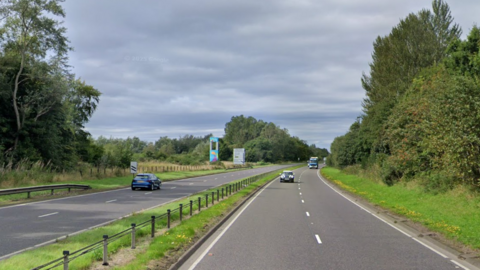 A dual carriageway separated by a central reservation covered in grass with a small fence. The road is surrounded by trees and grass. A couple of cars travel in both directions on the road.