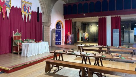 A view of a large room from the left hand corner. There are wooden benches lined up on the right and a small stage on the left, which has two chairs and a white table on it.