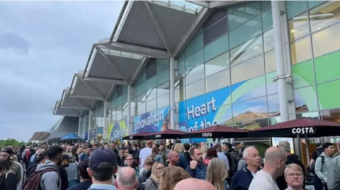 Along line of people outside Birmingham airport. There are four brown costa coffee umbrellas lined along the airport walls. 