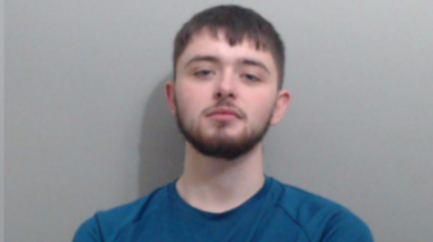 A man with brown hair and a beard. It is a police mugshot so he is staring impassively at the camera.