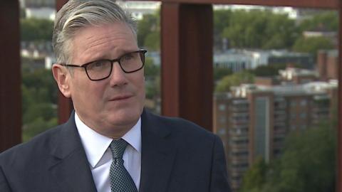 Sir Keir Starmer, who has swept back grey hair and is wearing black framed glasses, a dark suit over a white shirt and blue tie with dots. He is standing on a tall building and behind him is an iron framework and beyond that high-rise buildings and trees. 