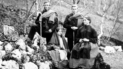 A black and white photograph showing a group of four people in the outdoors. A girl wearing a shawl and a woman wearing a coat and long striped dress are sitting on rocks. Behind them stand a man holding a shepherd's crook and a younger man with hands folded and resting on a stick. Both men have plaids over one shoulder.