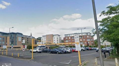 A carpark at Yorktown site in Sullivan Road, Camberley. There are numerous cars parked on the site and there are three blocks of flats on the side. There are also trees surrounding the carpark. 