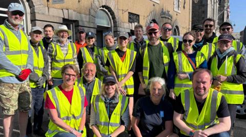 The photo shows a large group of volunteers wearing bright yellow high‑visibility vests, gathered together on a sunny day. Some of the volunteers are standing while others are crouching at the front. They are outside and behind them is a stone building with arched windows.