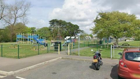 Google Street View image of Hayle Recreational Ground in Cornwall. A play park is in the background. A black motorbike and dark orange car are parked up outside the park's grounds. 