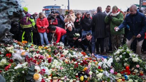 Magdeburg residents and emergency respondents in winter gear lay flowers and candles near the attack scene. At the forefront of the image are several bouquets of flower and plush toys and candles