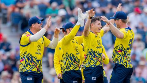 A group of Durham players congratulate Matthew Potts (second from right) on taking a wicket in a T20 Blast match in their yellow and navy blue T20 kit