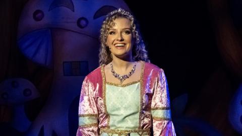 Alicia Belgarde wears a velvet pink and green dress. She has blonde curly hair and is wearing a tiara. She is stood on stage in front of a background which features mushrooms.