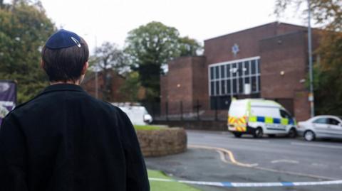 A young man stands looking away from the camera at a brick-built synagogue which is cordoned off by police tape. 