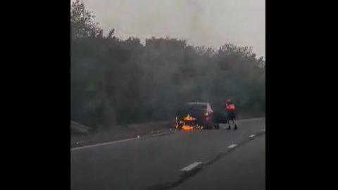Car on fire on the hard shoulder of the A12. The driver is seen getting out of the car. Flames can be seen. 