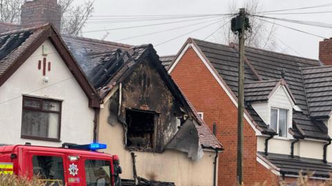 A semi-detached house with a roof burned exposing charred tiles and walls. There is a red fire truck with blue lights flashing.