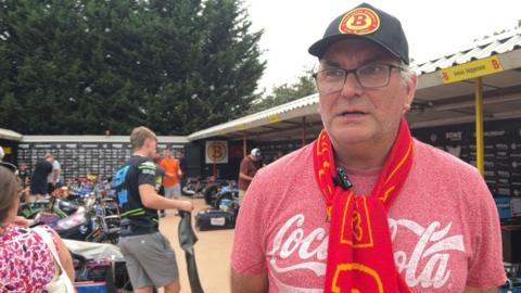 Dennis has a red and white t-shirt on that says "coca cola" he is wearing a yellow and red scarf around his neck, and a black cap with a red and yellow logo on it. He wears black glasses and is standing in front of a row of speedway bikes parked in a line.