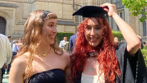 Grace and Caitlin stand in front of a university building together. Grace has long blonde hair and wears a black dress. Caitlin has died red hair and is wearing a graduation hat and gown.