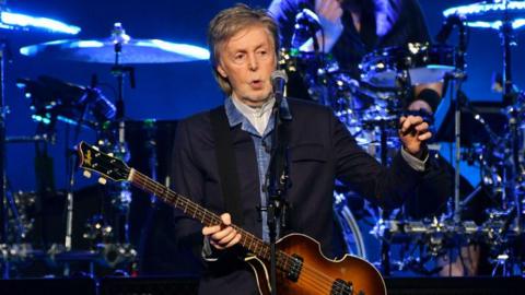 Sir Paul McCartney standing onstage. He has grey hair, and is wearing a dark blue jacket with light blue shirt. He is holding a bass guitar in his hands, with a drum kit visible in the background and a microphone in front of him. 