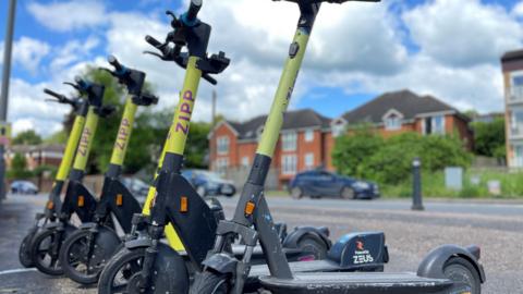 A row of five e-scooters with the yellow and purple 'Zipp' logo on them, stationary at the side of a residential street with trees and houses in soft focus behind them