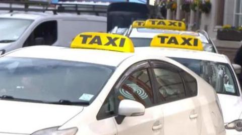 A picture of a line of taxis in St Helier. They are white with yellow and black taxi signs on their roofs.