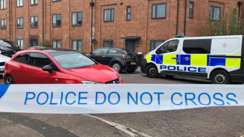 A close-up shot of police tape saying 'Police do not cross' with cars and a police van in the background, parked on a residential street