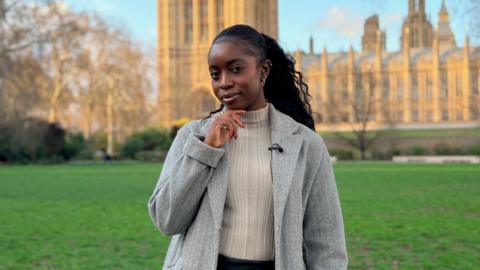 Jonelle Awomoyi looks at the camera resting her chin on her hand in front of Houses of Parliament. She wears a slick ponytail, a beige jumper and a grey coat.