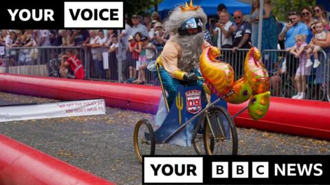A competitor in Castle Douglas Soapbox Derby. A man dressed as a mermaid is riding a three-wheeled bike along the course. A crowd of spectators is watching from behind a barrier.