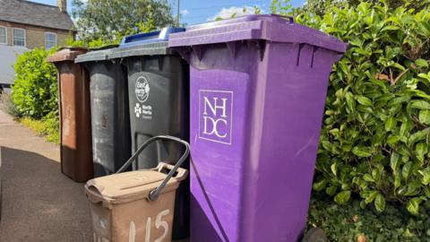 Four wheelie bins are lined up against a hedge. One is purple, two are black, and one is brown. They are variously branded North Herts District Council and East Herts Council. Standing in front of them is a much smaller brown food waste caddy with a handle and the number "115" hand-painted on it.