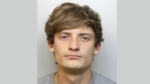 Police mugshot of Harry Charlton. He is wearing a grey t-shirt and standing against a grey background, looking at the camera. He has green-grey eyes and shaggy, light brown hair which is sweeping across his forehead.