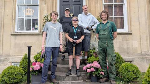 Five people stand in front of a Georgian-style house, on the steps leading up to it. There are flower pots and small hedges going upwards to it.