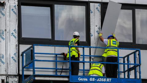 Three workmen in hi-vis jackets and helmets removing cladding from a building