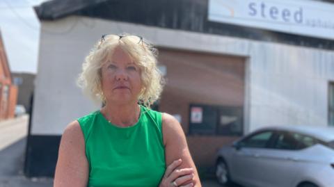 A woman with crossed arms looking irked outside a factory building