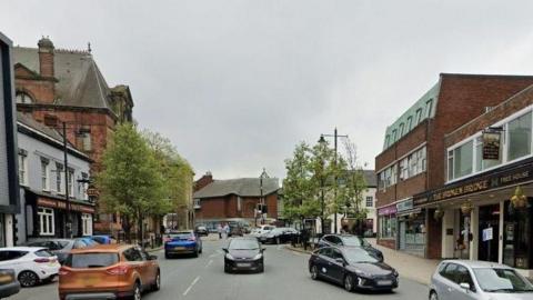 A busy street with shops and pubs at either side, and several cars driving up and down. The sky is grey.