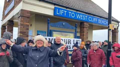 A crowd of people dressed in waterproof coats and many with hats on stand in front of a small building on a beach promenade. There is a blue sign on the building saying, lift to West Cliff beach. The crowd has put a banner up across the building but it cannot be read as they stand in front if it.