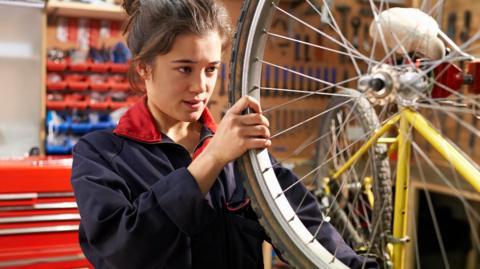 A young woman with brown hair tied back in a bun is wearing a navy overall with a red collar. She is in a workshop and has a yellow bike attached to a stand in front of her, and is turning the back wheel with her hands. There are lots of tools hanging up on the wall behind her.