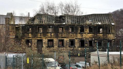 A stone built mill building with empty windows and an exposed roof with collapsed beams