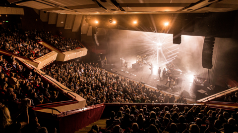 A general view picture of a band playing at the Hexagon with bright lights illuminated on the stage.