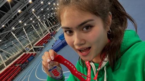 Athlete Issey Kyson holds up a medal while standing on an indoor race track. She has long brown hair and wears a green hoodie