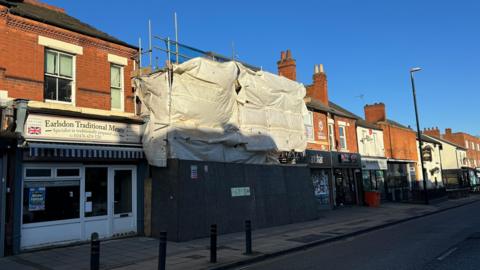 A building is covered by scaffolding which itself has white plastic sheeting covering it. At the bottom half of the building, dark grey hoardings surround the property footprint, which is in the middle of the street.