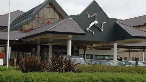 The front entrance of Noble's Hospital on the Isle of Man, which features slanted roofs and a large metal three-legs-of-Man on the front. Several cars and a van are parked in front of the entrance, obscured partially by a green hedgerow.