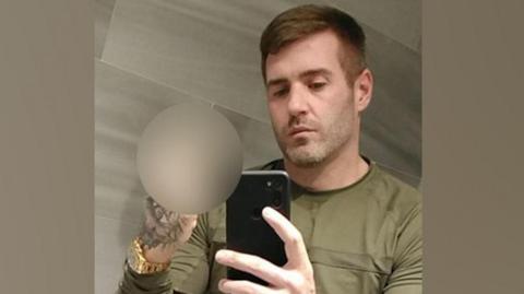Jamie Rothwell is posing for a selfie in a mirror. He is wearing a khaki top and a gold watch. He is making an offensive gesture with his right hand.