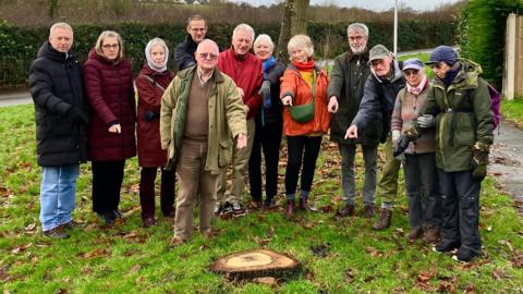 Meole residents standing on a grassy verge in front of a small oak stump, looking unhappy, with a sweet chestnut tree and hedgerow behind them. They wear jackets and some have headwear including caps and scarves. Many are pointing at the stump.