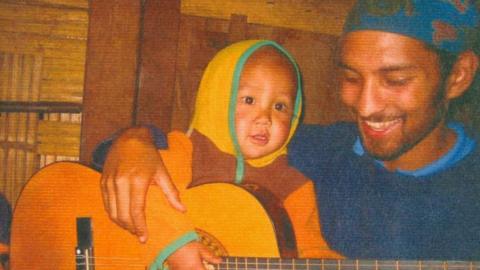 Alex Ratnasothy dressed in a blue bandana and jumper with a child on his lap who he is helping play guitar while travelling in Burma.
