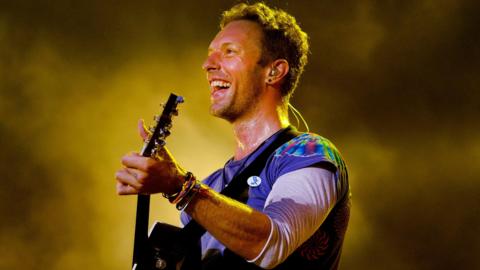 Chris Martin smiling while performing on stage and holding a guitar. He has short dark hair and is wearing a multicoloured T-shirt with long arms. He is smiling and behind him is dry ice smoke lit by a yellow light.