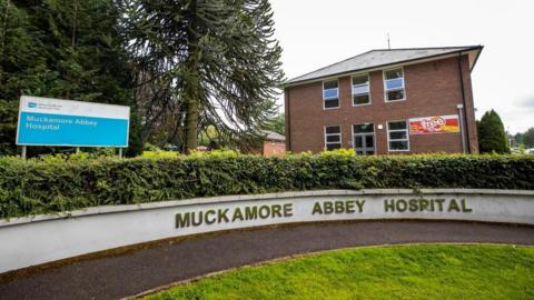 A sign that says Muckamore Abbey Hospital in gold writing on a white wall. 