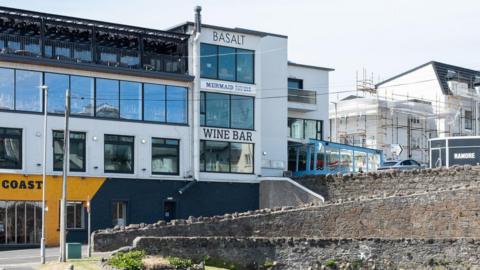 The exterior of the Ramore restaurants building in Portrush. The image shows a tall four storey building with an eatery on each floor. They are marked out by signage, including signs for Coast, Wine Bar, mermaid and Basalt. There is a black trailer with Ramore written on it at the top of the hill.