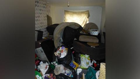 Room in flat shows light bulb with no lamp shade, bags of rubbish all over the floor and furniture in a piled with a dirty white sheet covering the window 