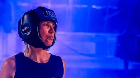 Teresa wearing a blue sleeveless boxing jersey and a blue helmet standing inside the boxing ring with blue lights. 